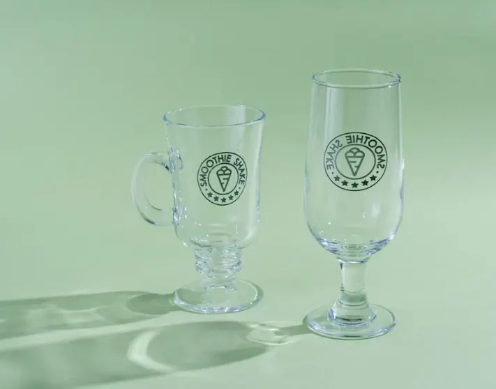Favism3D: Servicio de grabado láser en vidrio y cristal para personalización de vasos, copas y regalos promocionales. Dos vasos de cristal con logo 'SMOOTHIE SHAKE' grabado con láser, mostrando personalización en vidrio.