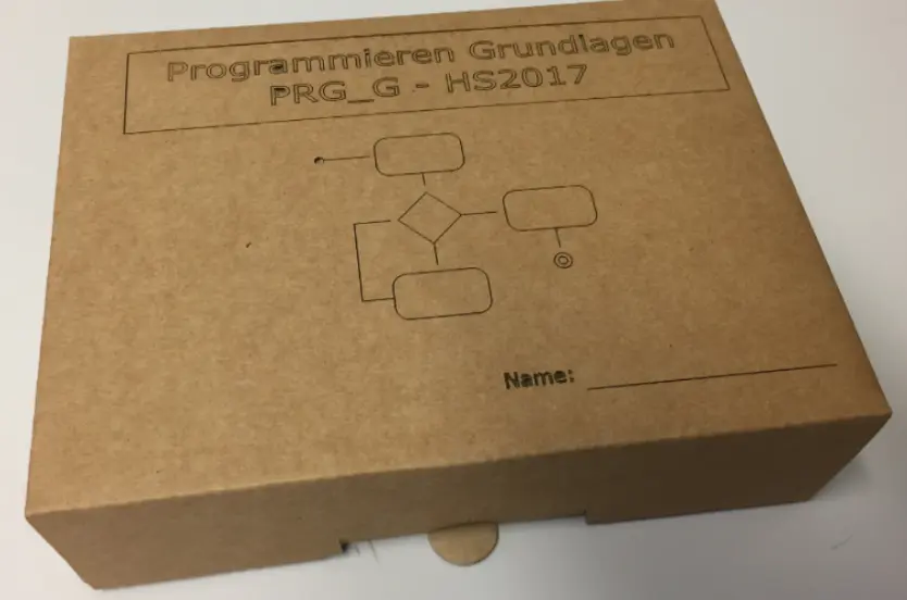 Favism3D: Grabado y corte láser en cartón y cartulina para packaging personalizado, eventos y material didáctico. Caja de cartón kraft grabada con láser, mostrando texto 'Programmieren Grundlagen' y un diagrama de flujo, ideal para packaging.