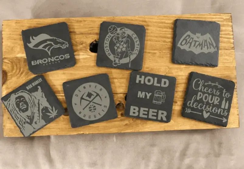 Favism3D: Grabado láser en piedra y pizarra para posavasos, regalos personalizados y decoración única. Conjunto de siete posavasos de pizarra negra grabados con láser, mostrando diversos logos y frases personalizadas.
