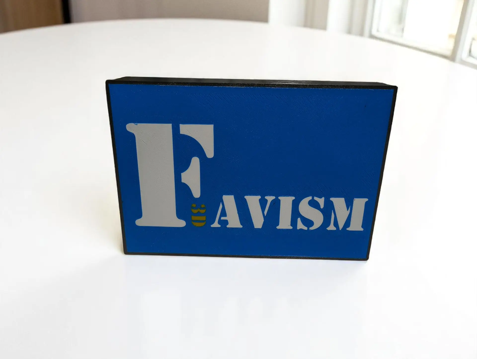 Letreros y carteles 3D personalizados, señalética de eventos, decoración de mesa, rótulos impresos, favismo, diseño a medida, Málaga. Letrero de mesa personalizado impreso en 3D con la letra 'F' y el texto 'FAVISM' en gris sobre fondo azul, ideal para eventos o negocios.